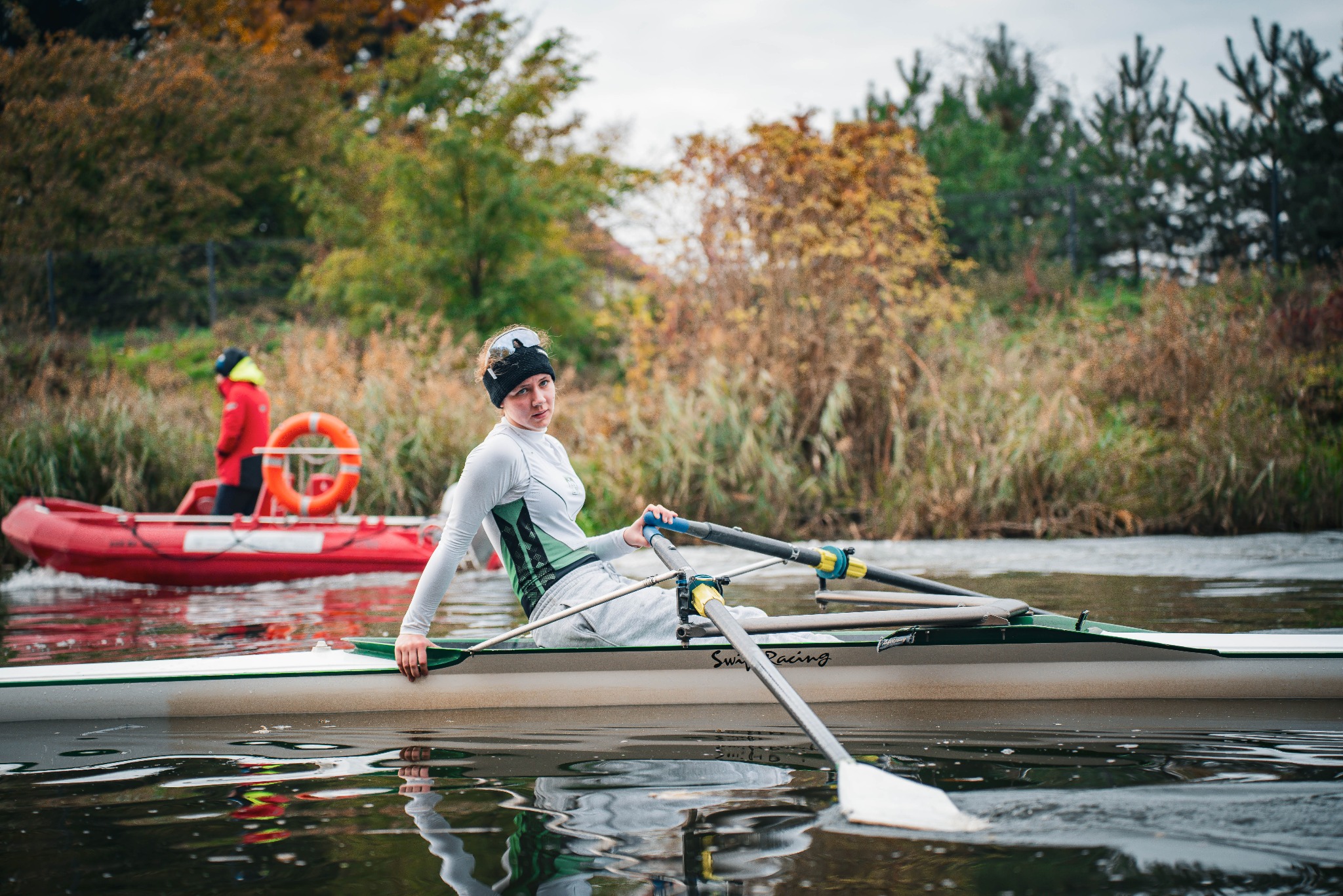 Kobieta w łodzi wioślarskiej, na wodzie, w tle drzewa i osoba w czerwonej kurtce w pontonie. Ujęcie z bliska, widać wiosła i refleksje w wodzie.