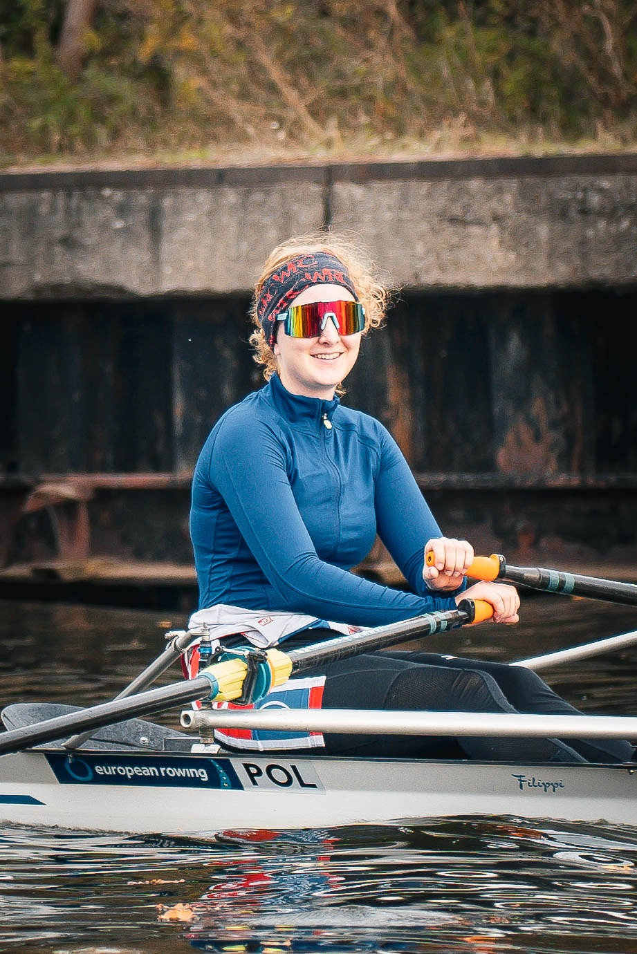 Uśmiechnięta wioślarka w okularach przeciwsłonecznych i opasce na głowie, wiosłuje w łodzi z napisem 'POL' na tle betonowego nabrzeża.