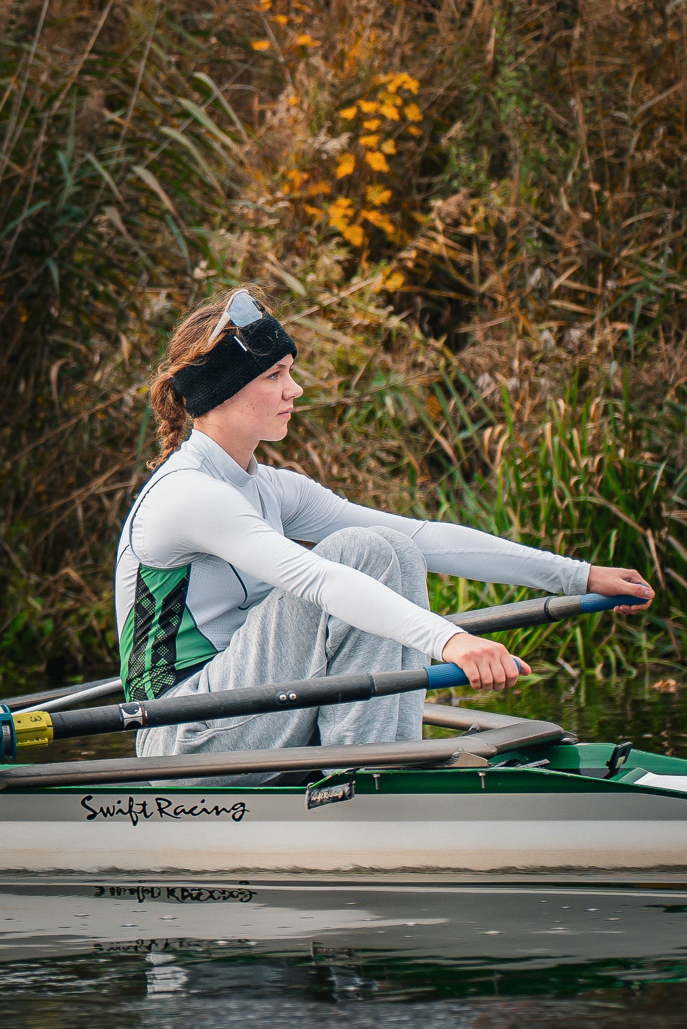 Młoda kobieta w trakcie wiosłowania na wodzie, ubrana w sportowy strój, z wiosłami w dłoniach. W tle jesienna roślinność i spokojna woda.