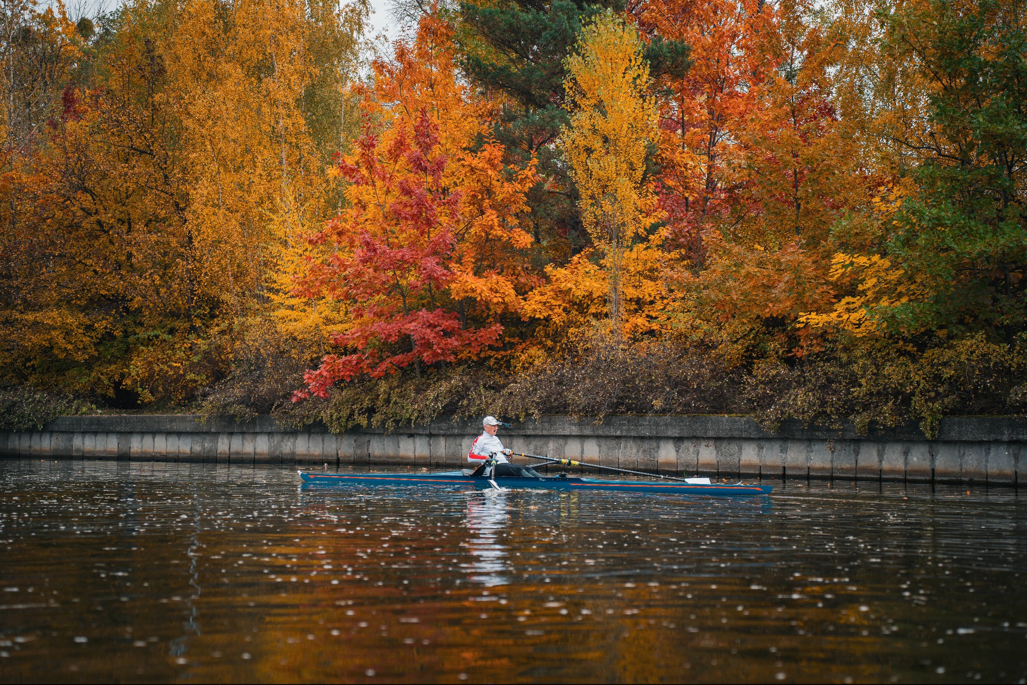 Wioślarz na jeziorze w jesiennej scenerii. Łódź sunie po wodzie, otoczona drzewami w odcieniach czerwieni, pomarańczy i żółci. Scena uchwycona w naturalnym świetle.