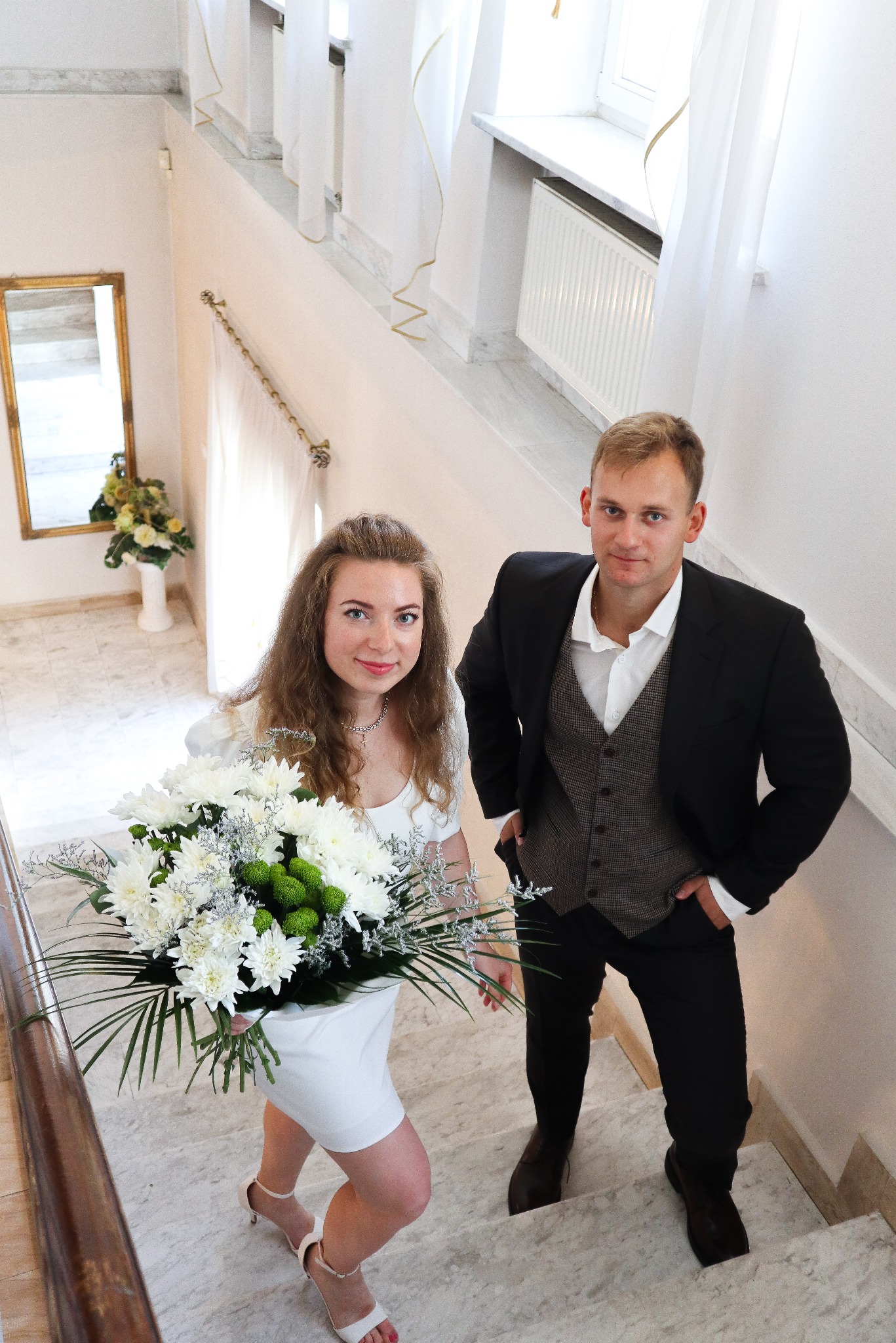 Para młoda na marmurowych schodach w jasnym wnętrzu. Panna młoda trzyma bukiet białych kwiatów. Widok z góry.