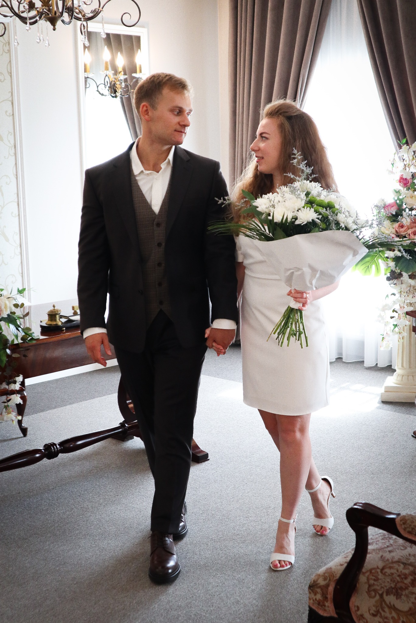 Uśmiechnięta para młoda, on w garniturze, ona w białej sukience z bukietem, idąca w jasnym wnętrzu. Kadr z ceremonii ślubnej, wzajemne spojrzenia.
