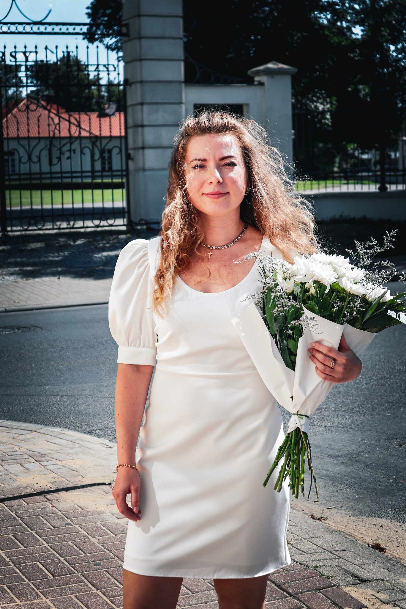 Uśmiechnięta kobieta w białej sukience trzyma bukiet białych kwiatów na tle bramy z kutego żelaza i budynku z czerwoną dachówką.