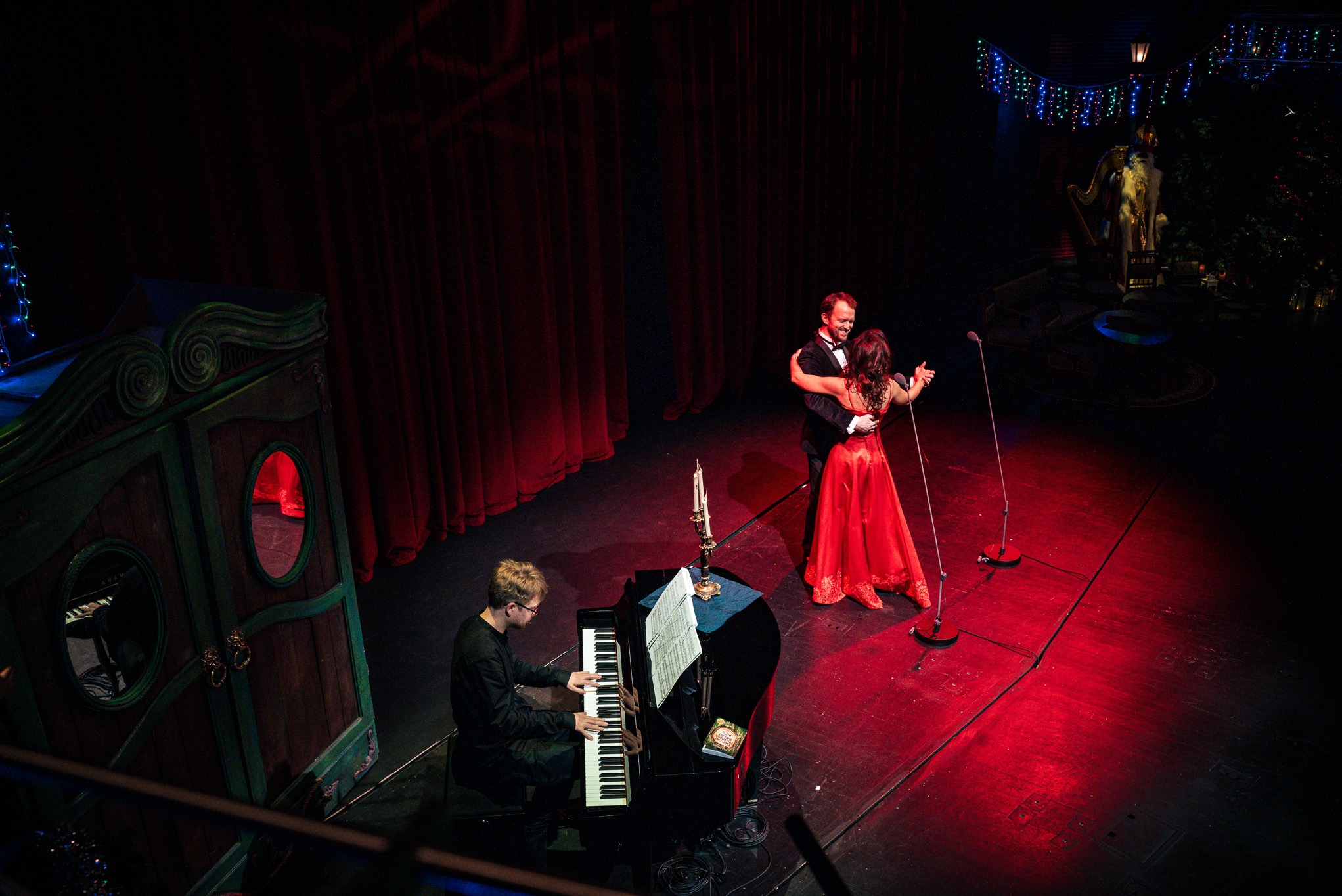 Para tańczy na scenie przy akompaniamencie pianisty. Czerwone światła, dekoracje świąteczne i ozdobna garderoba tworzą elegancką atmosferę wydarzenia.