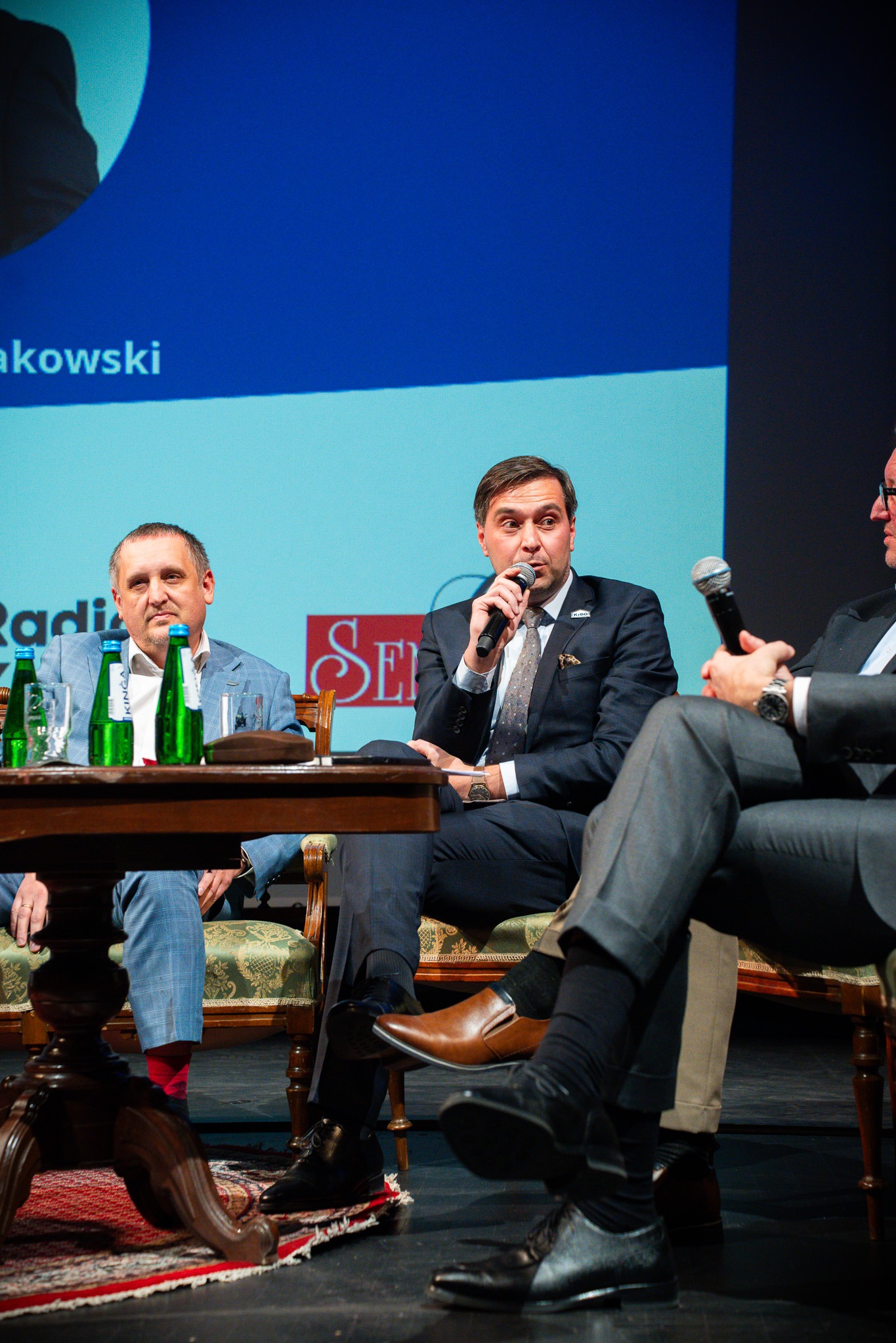 Panel dyskusyjny w Warszawie: Mężczyzna w garniturze przemawia do mikrofonu, siedząc obok innych prelegentów. Scena, oświetlenie eventowe, profesjonalizm.