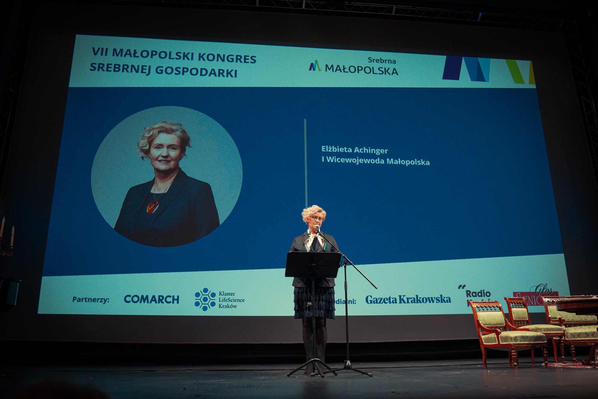 Prelegentka na scenie podczas VII Małopolskiego Kongresu Srebrnej Gospodarki. W tle ekran z logiem i zdjęciem prelegentki Elżbiety A.
