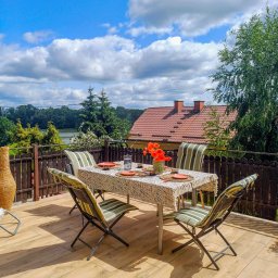 Dom Milikowo Ewa Lechocka - Umeblowany balkon z drewnianą podłogą i widokiem na jezioro. Stół nakryty do posiłku, krzesła w paski. Słoneczny dzień, błękitne niebo z chmurami.