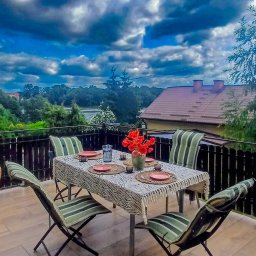 Dom Milikowo Ewa Lechocka - Balkon z nakrytym stołem, krzesłami i widokiem na krajobraz z budynkiem. Dekoracja stołu obejmuje czerwone kwiaty i naczynia.