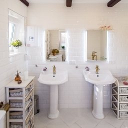 Dom Milikowo Ewa Lechocka - Jasna łazienka z dwoma umywalkami na postumentach, białe płytki na ścianach i podłodze, drewniane belki na suficie, lustra z podświetleniem i dekoracje.