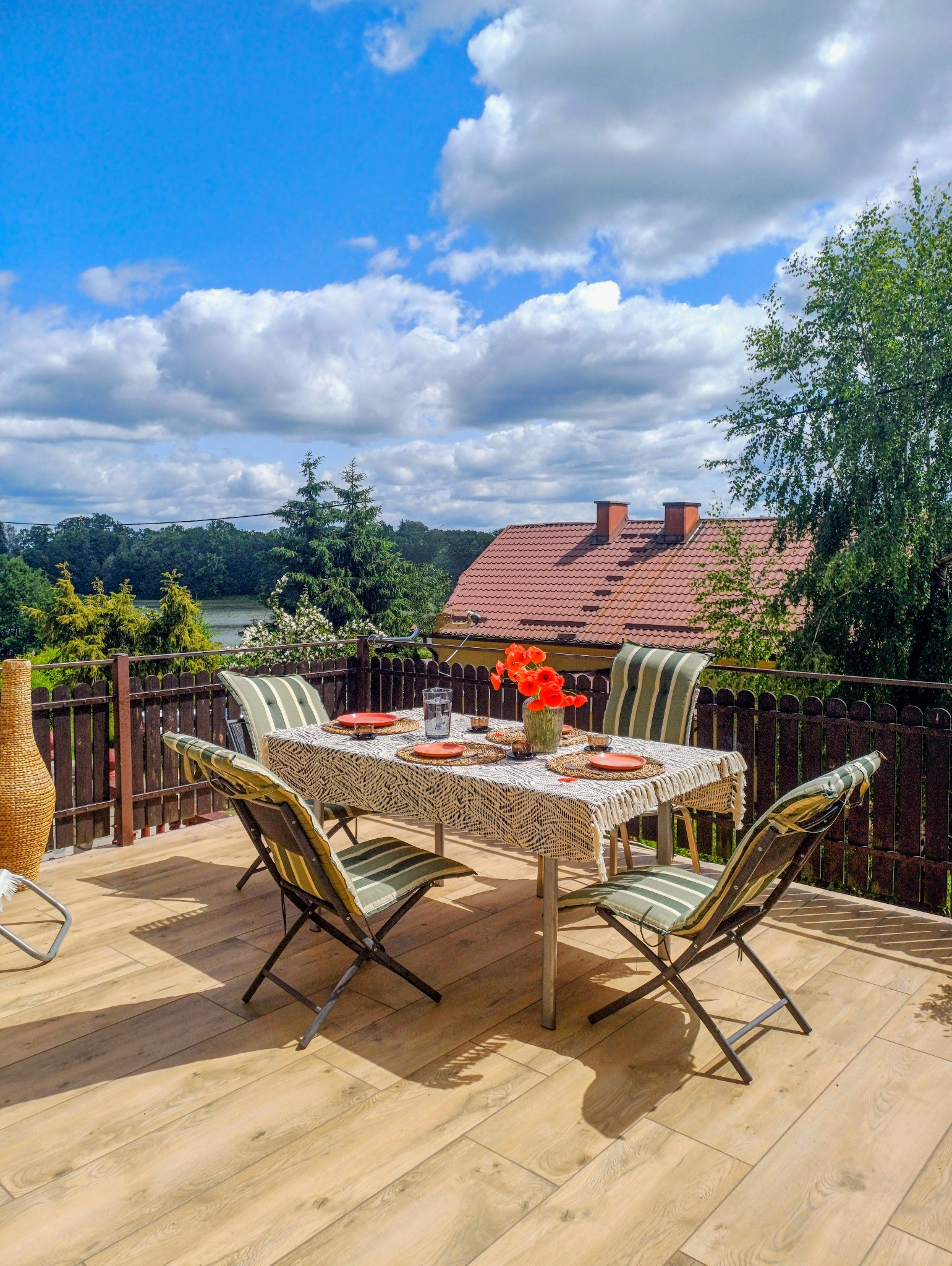 Umeblowany balkon z drewnianą podłogą i widokiem na jezioro. Stół nakryty do posiłku, krzesła w paski. Słoneczny dzień, błękitne niebo z chmurami.