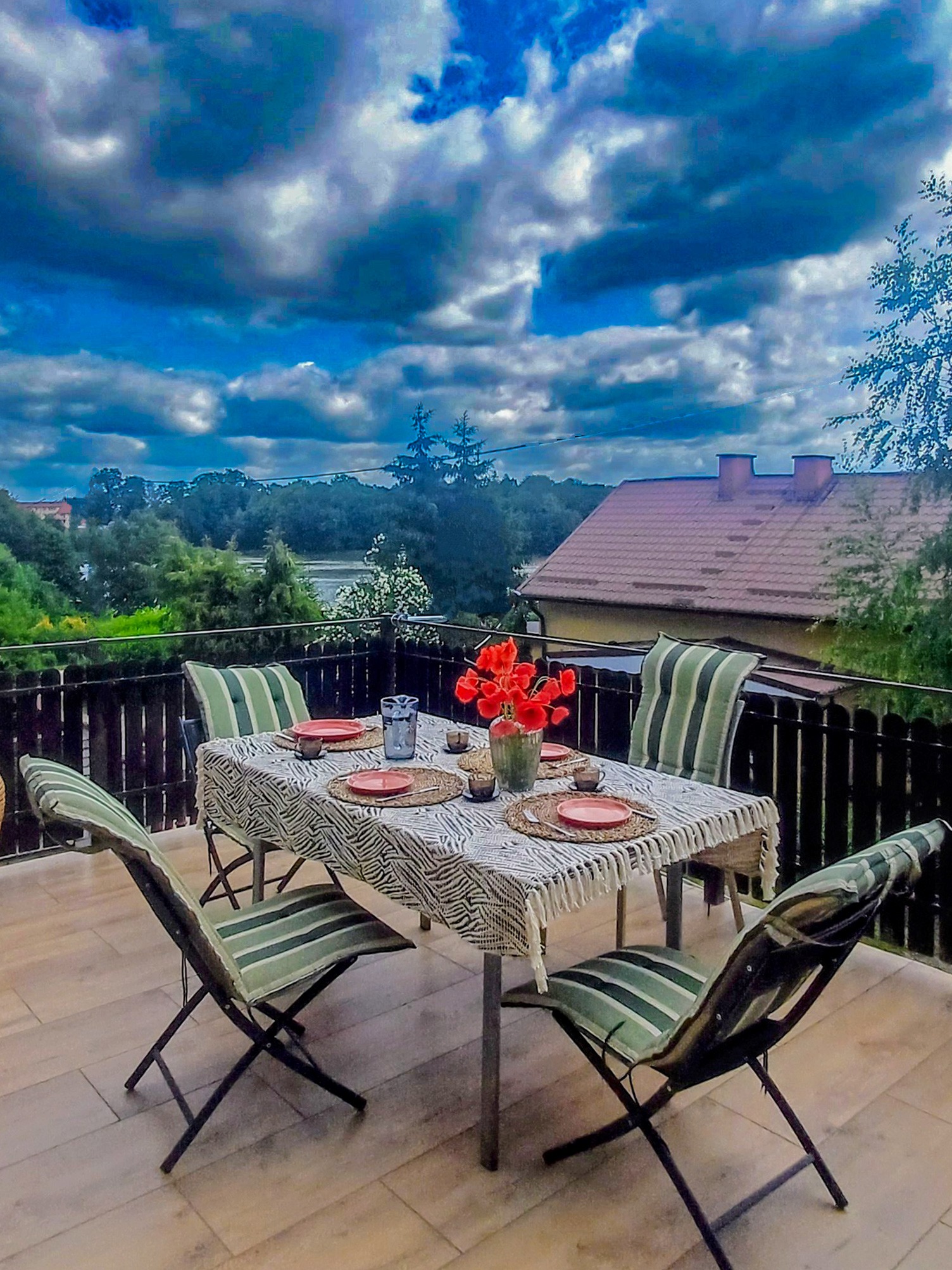 Balkon z nakrytym stołem, krzesłami i widokiem na krajobraz z budynkiem. Dekoracja stołu obejmuje czerwone kwiaty i naczynia.