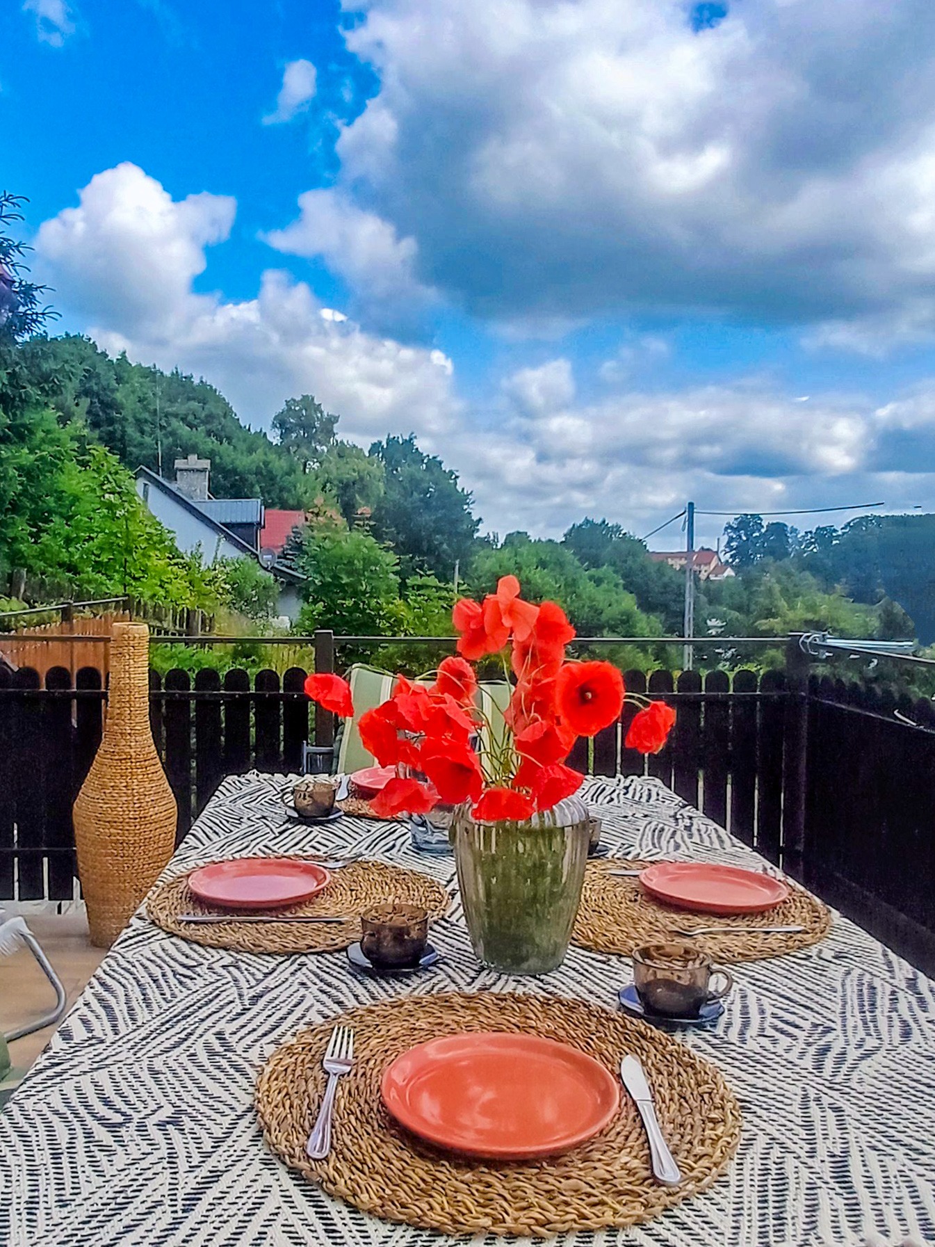 Ustawiony stół na balkonie z widokiem na zieleń i niebo. Czerwone maki w wazonie dodają koloru.
