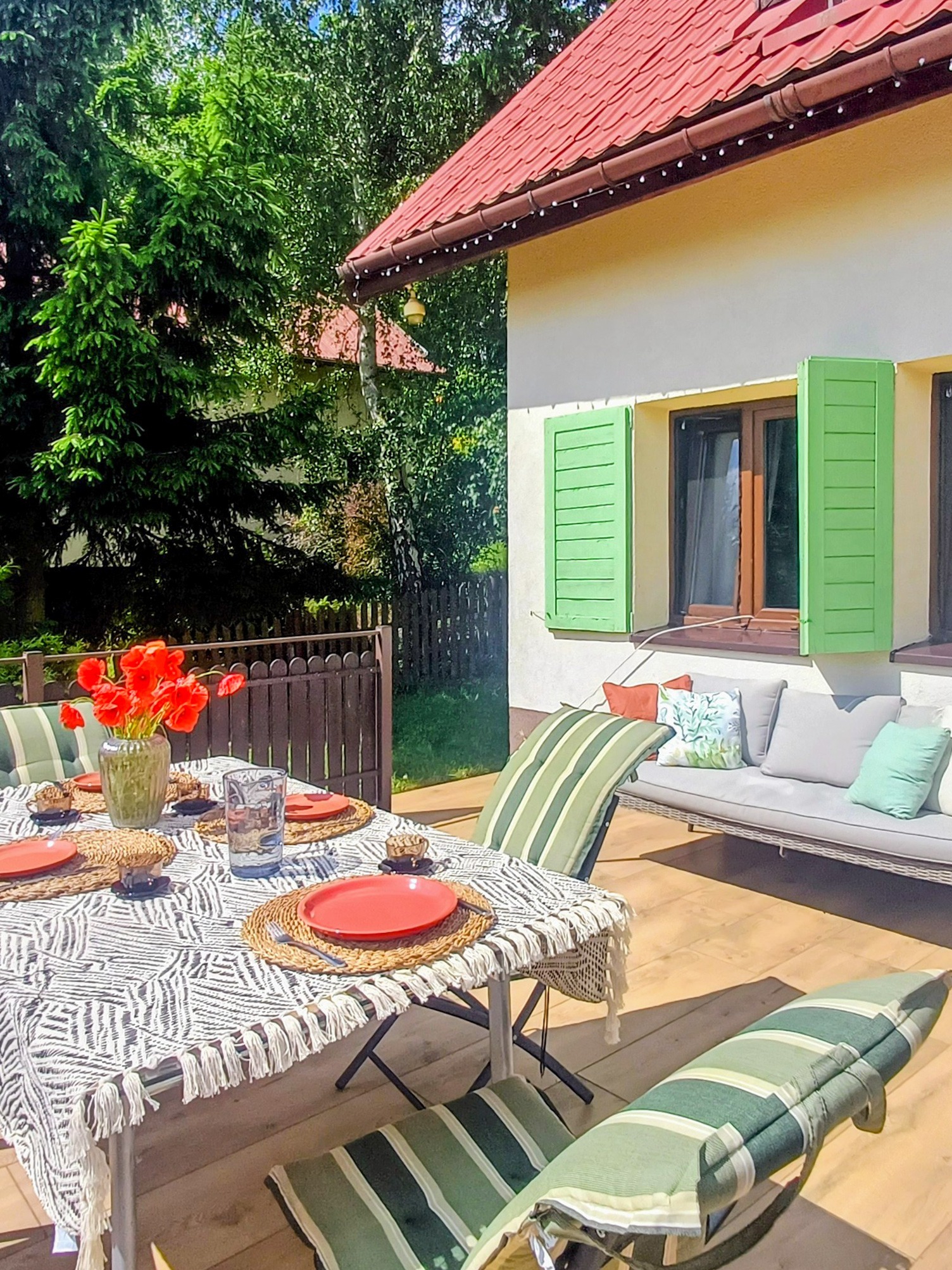 Letni taras z nakrytym stołem, czerwone maki w wazonie, zielone okiennice, sofa z poduszkami. Słoneczny dzień, relaks na świeżym powietrzu.