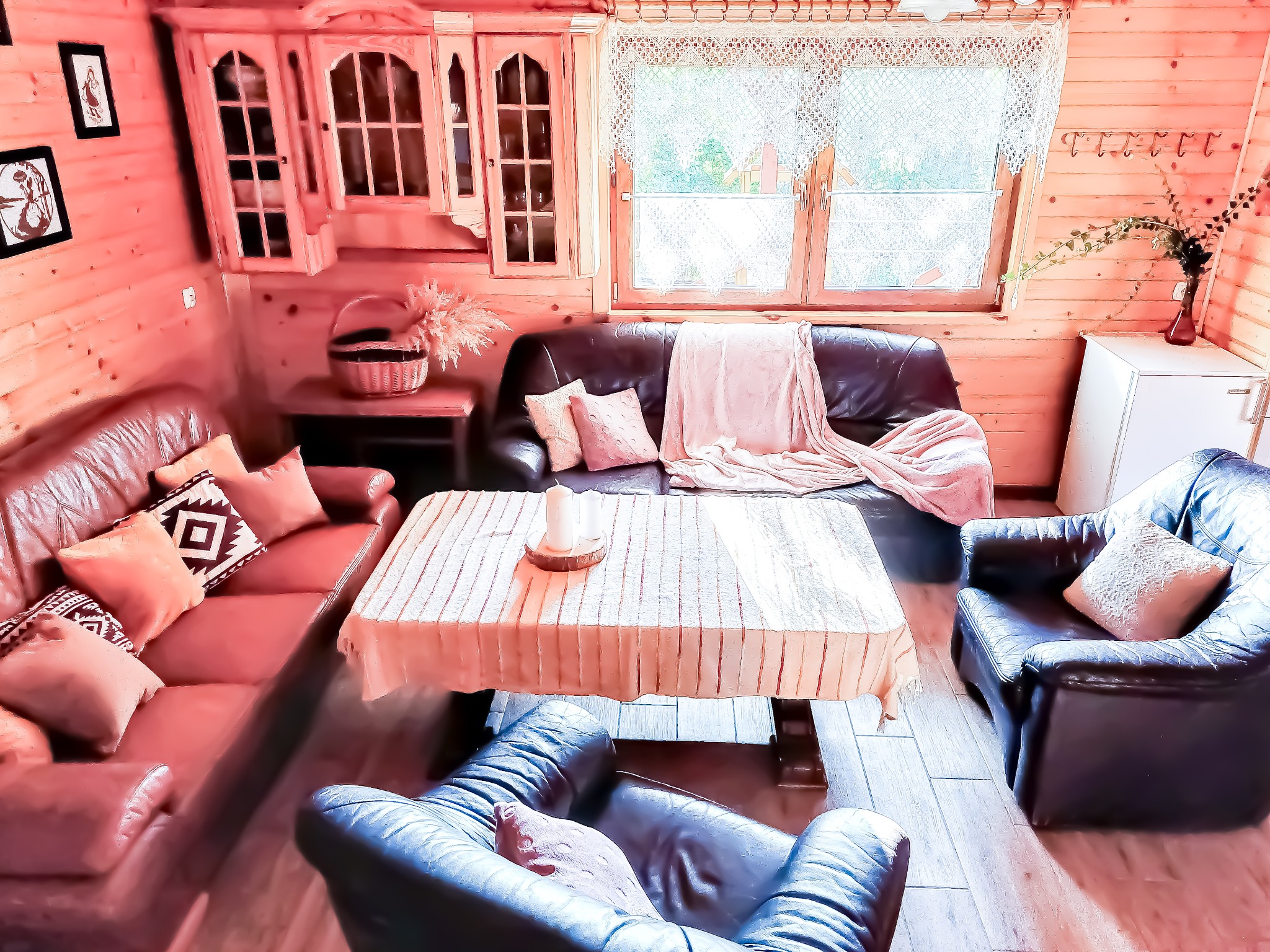 Przytulne wnętrze drewnianego domu z sofami, fotelami i stołem nakrytym obrusem. Na stole świece, w tle okno z koronkową firanką.