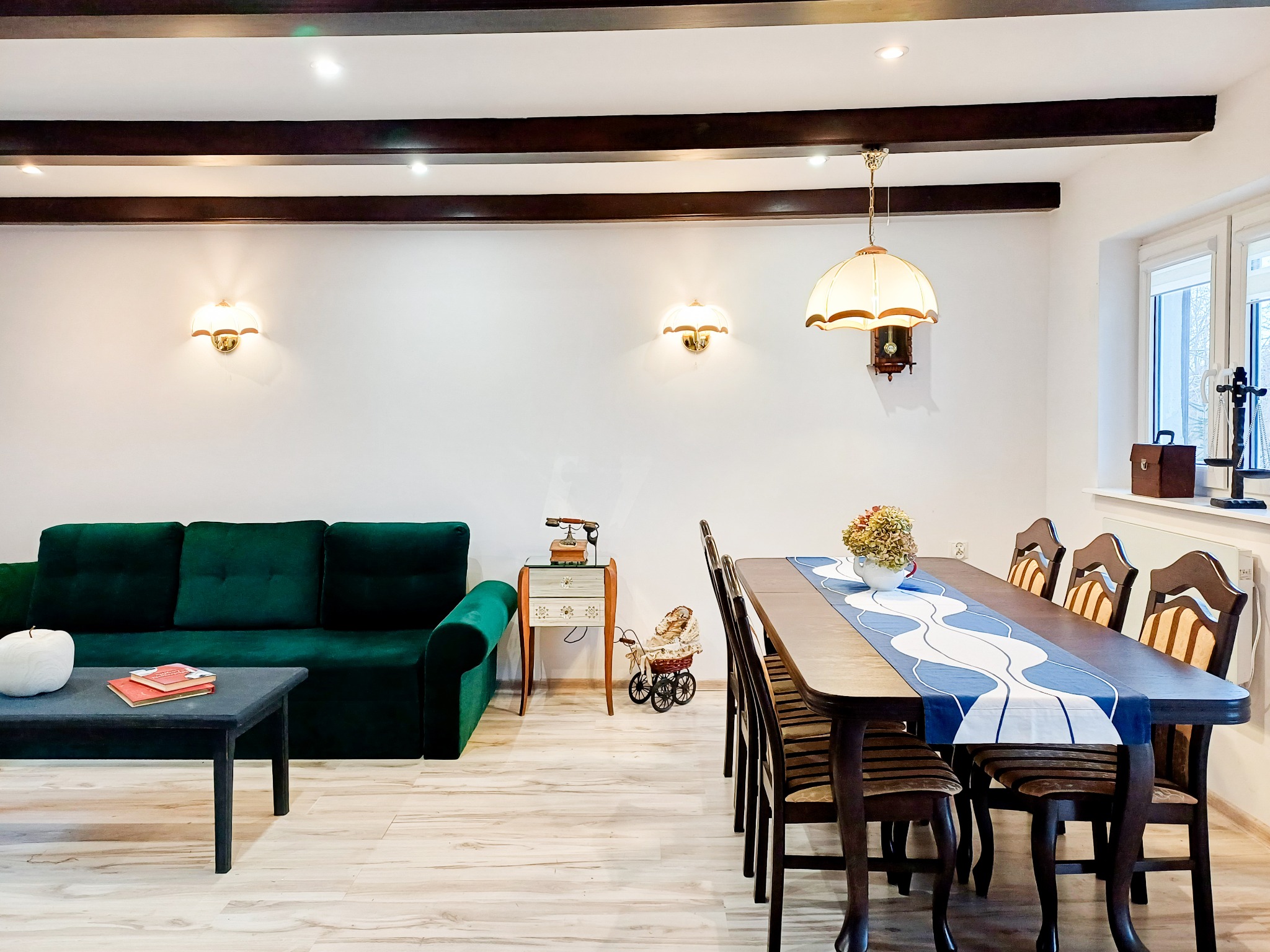 Jasne wnętrze salonu z drewnianymi belkami sufitowymi, zieloną sofą i stołem z dekoracją, oświetlone lampami ściennymi i wiszącą lampą z zegarem.