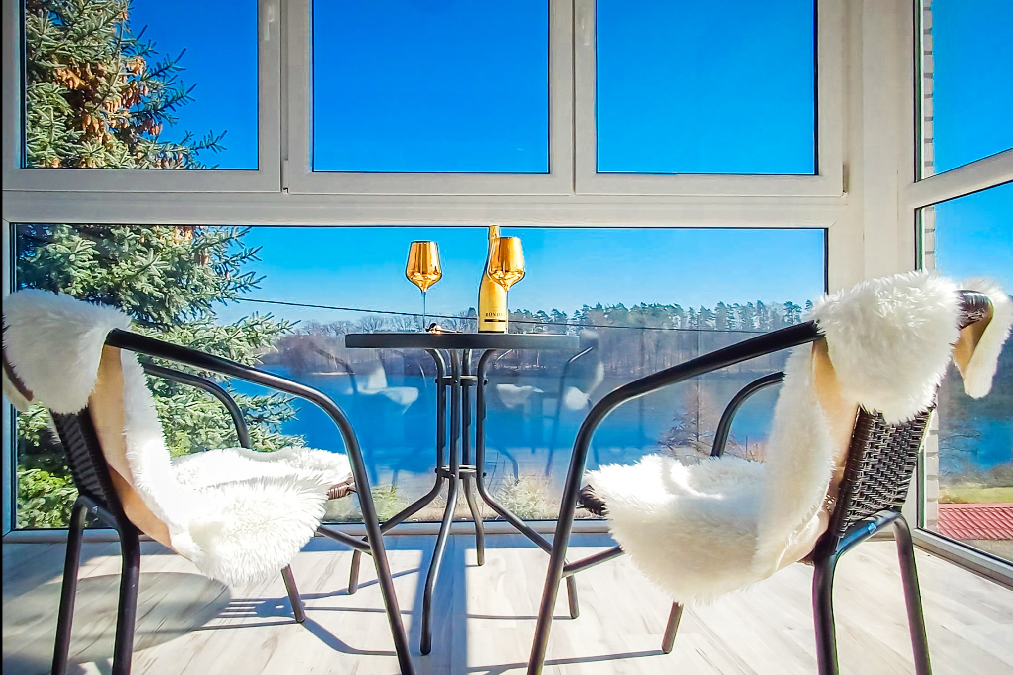 Słoneczny balkon z widokiem na jezioro, dwa krzesła z białymi poduszkami, stolik z butelką i kieliszkami. Relaksacyjny nastrój, idealne miejsce na odpoczynek.