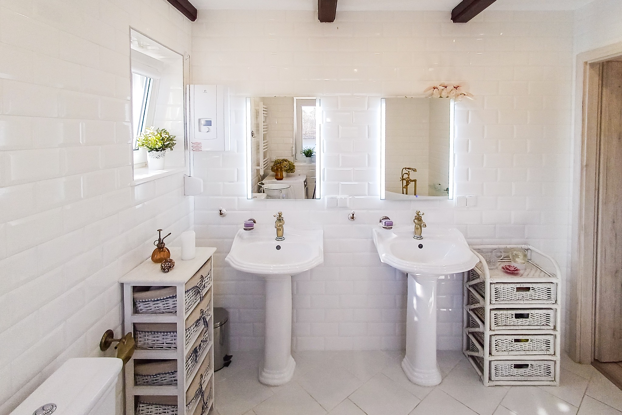 Jasna łazienka z dwoma umywalkami na postumentach, białe płytki na ścianach i podłodze, drewniane belki na suficie, lustra z podświetleniem i dekoracje.