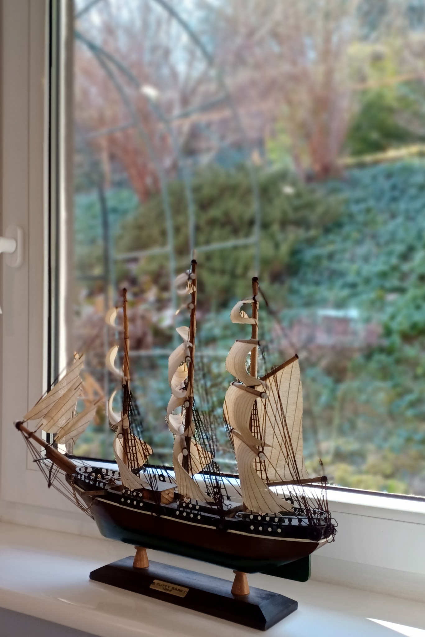 Model żaglowca Cutty Sark na parapecie okna z widokiem na zieleń. Detaliczne olinowanie, kremowe żagle i mahoniowy kadłub. Dekoracja wnętrza w neutralnym oświetleniu.