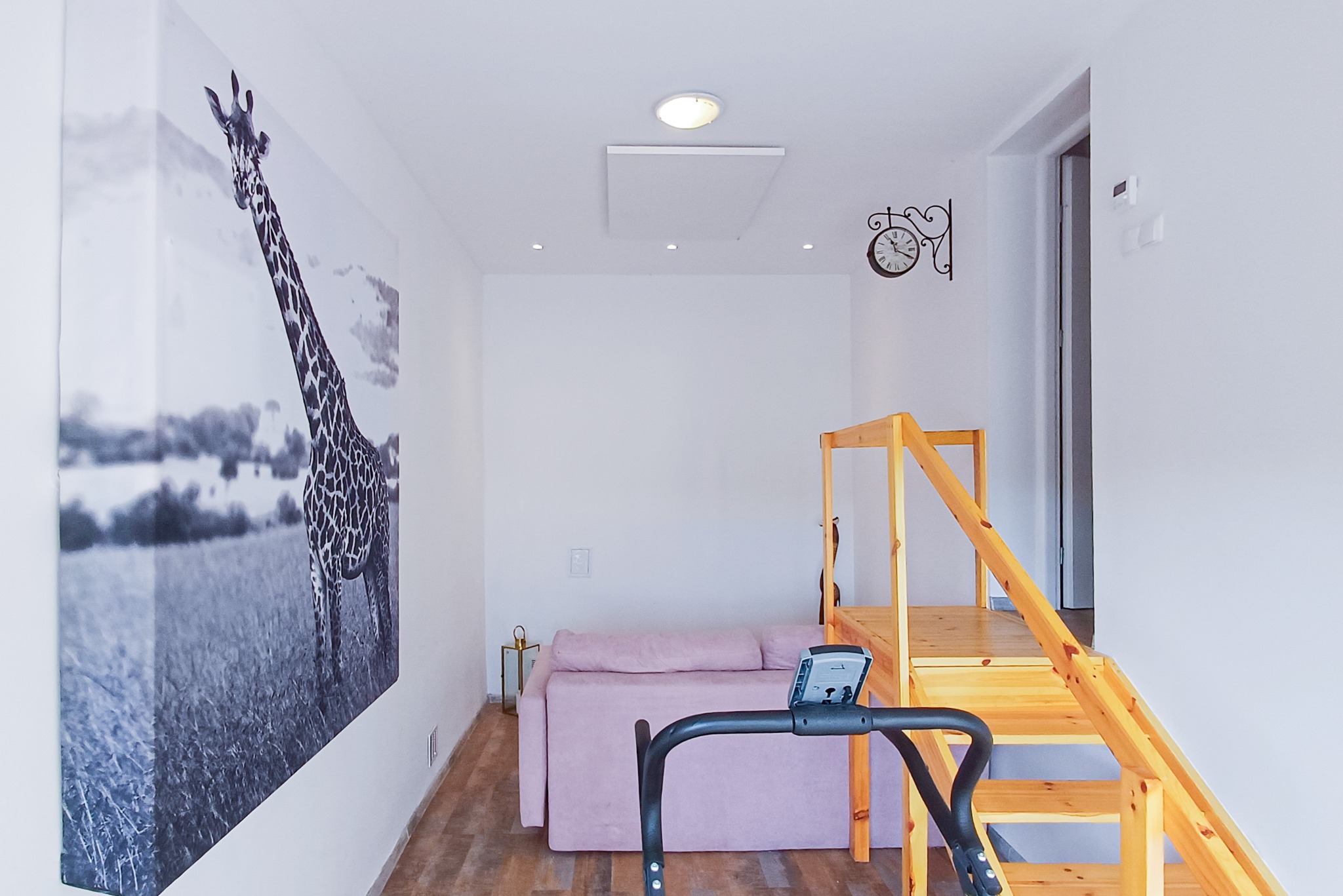 Jasne wnętrze z drewnianymi schodami, sofą i bieżnią; na ścianie duży obraz z żyrafą. Oświetlenie sufitowe i dekoracyjny zegar.
