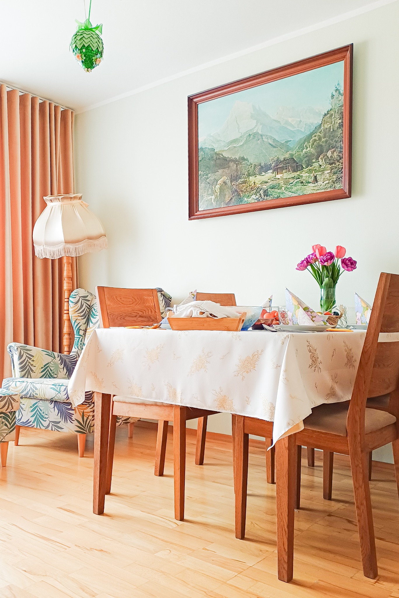 Jasne wnętrze jadalni z drewnianym stołem nakrytym białym obrusem w kwiaty, krzesłami i bukietem tulipanów. Na ścianie obraz z górskim pejzażem.