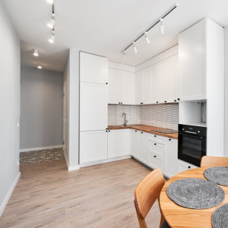 Jasna kuchnia po remoncie: białe szafki z drewnianym blatem i czarnymi uchwytami, drewniana podłoga, oświetlenie punktowe, okrągły stół z krzesłami.