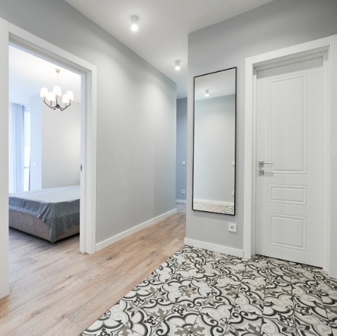 Wykończony korytarz we Wrocławiu z lustrem w czarnej ramie i wzorzystą posadzką. Widoczne drzwi i wejście do pokoju z łóżkiem. Ściany w odcieniach szarości.