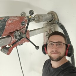 DOMIDOM Przemysław Insiński - Elektryk w okularach i słuchawkach ochronnych, obok wiertnica Hilti DD 120-U podczas wiercenia w ścianie. Praca przy instalacji elektrycznej.
