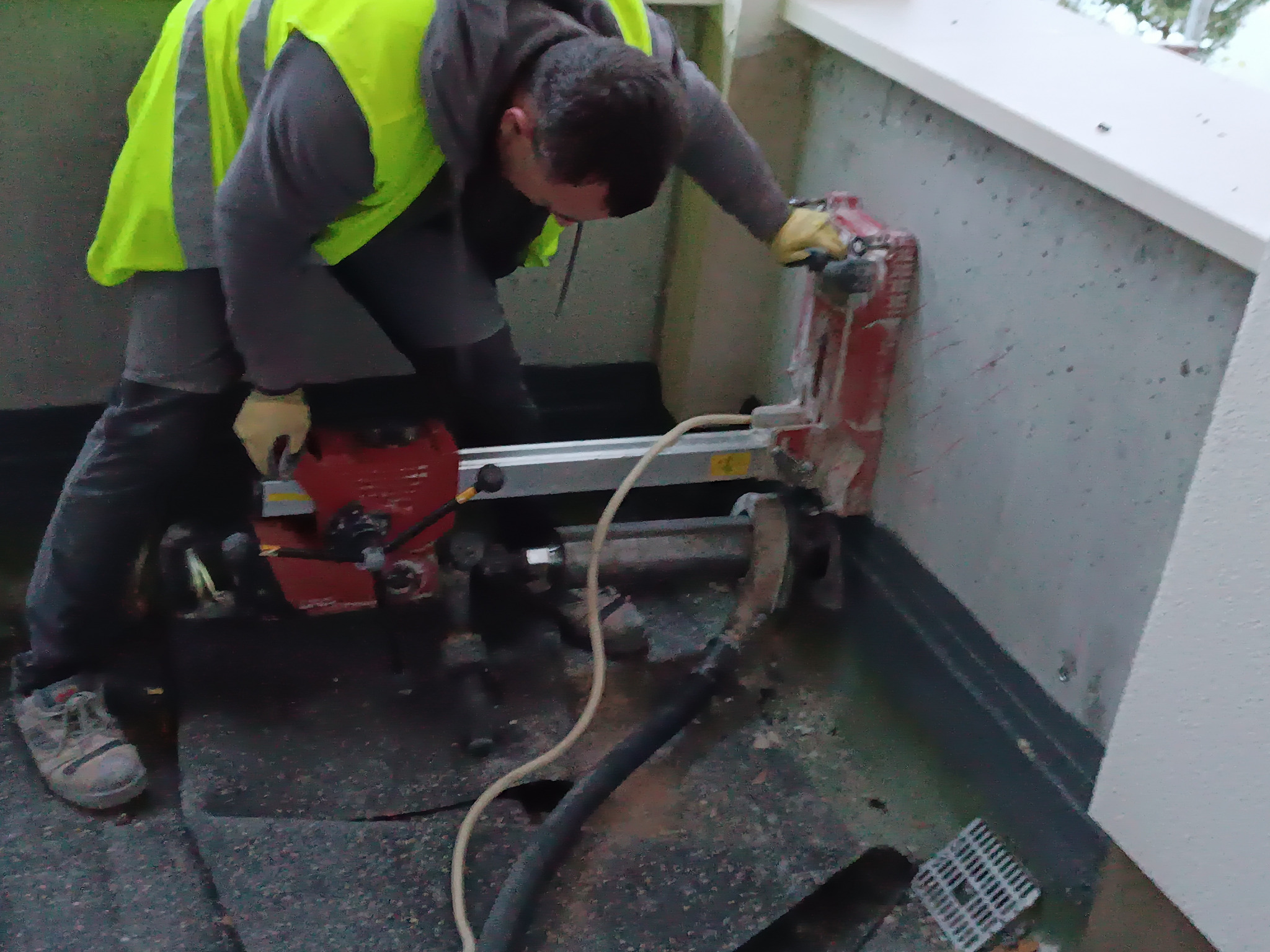 Pracownik w kamizelce wierci otwór w ścianie za pomocą wiertnicy rdzeniowej. Widoczne przewody i osprzęt. Praca wykonywana na zewnątrz budynku.