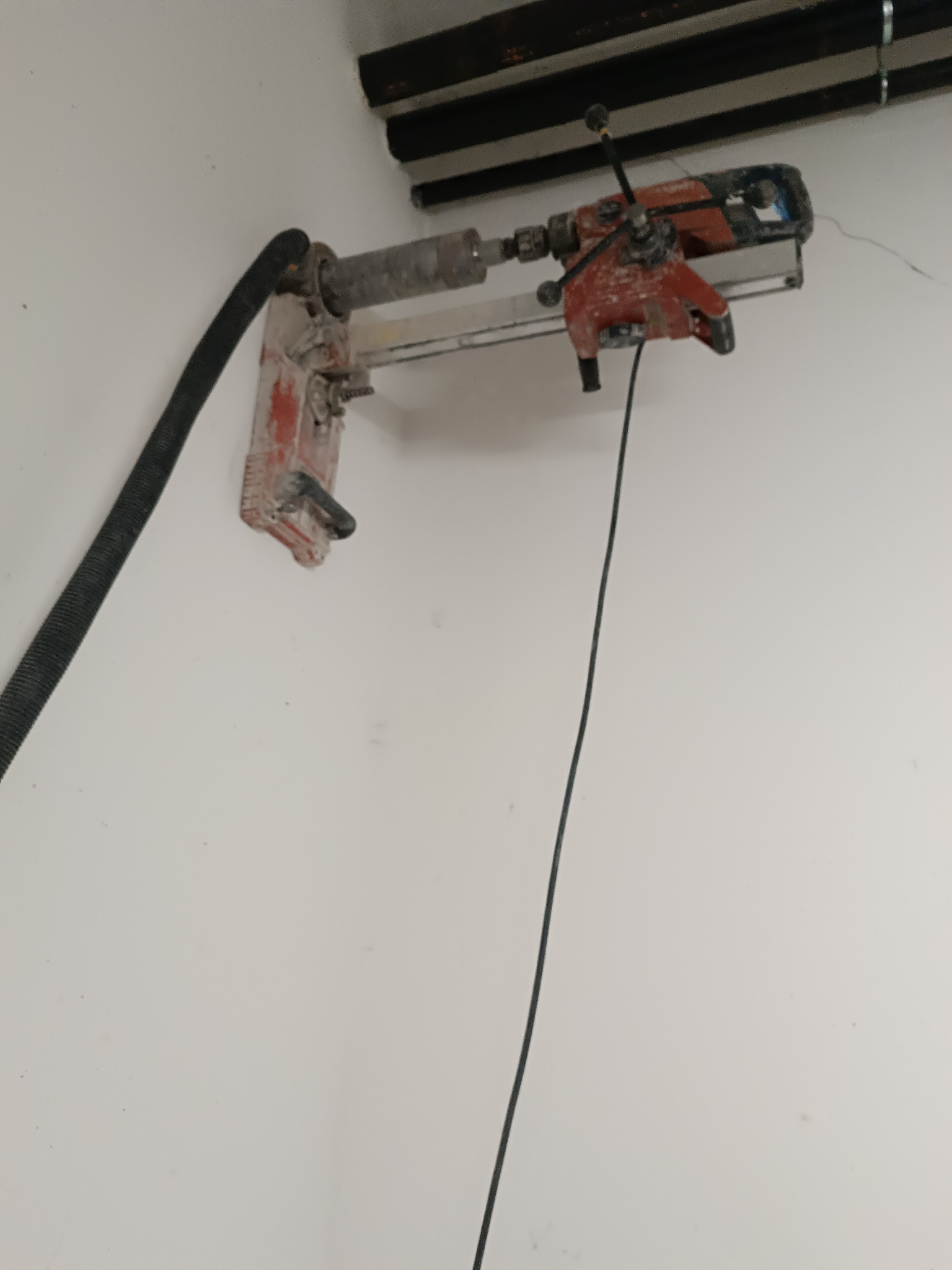 Wiertnica diamentowa przymocowana do ściany, z podłączonym odkurzaczem. Widoczne ślady wiercenia i przewód zasilający. Ujęcie z bliska, z boku.