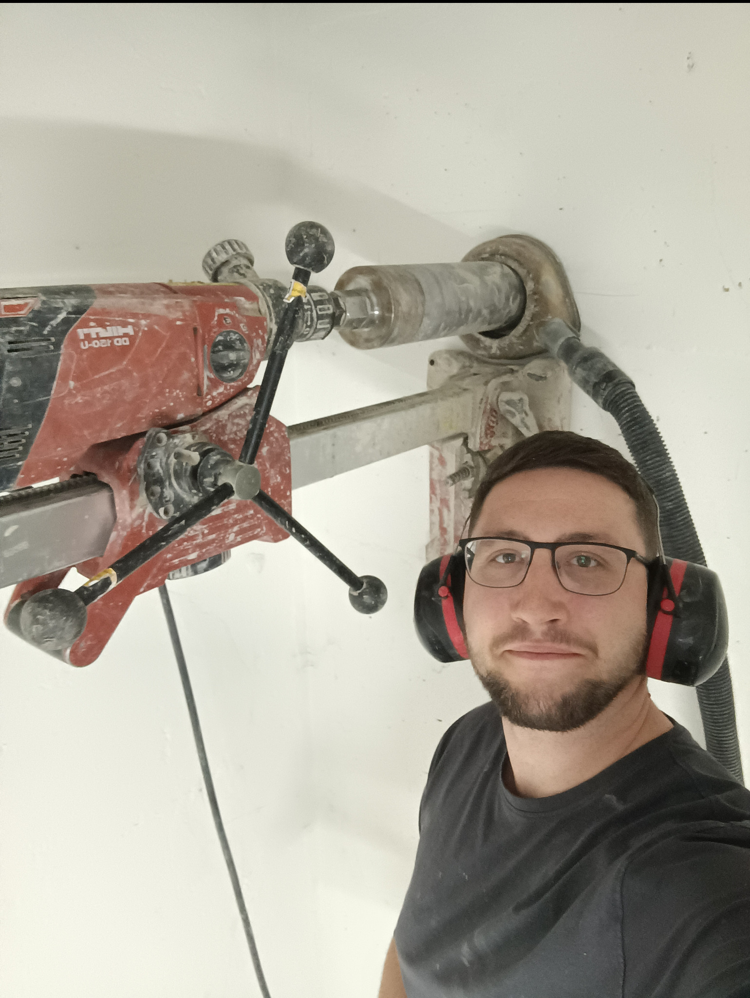 Elektryk w okularach i słuchawkach ochronnych, obok wiertnica Hilti DD 120-U podczas wiercenia w ścianie. Praca przy instalacji elektrycznej.