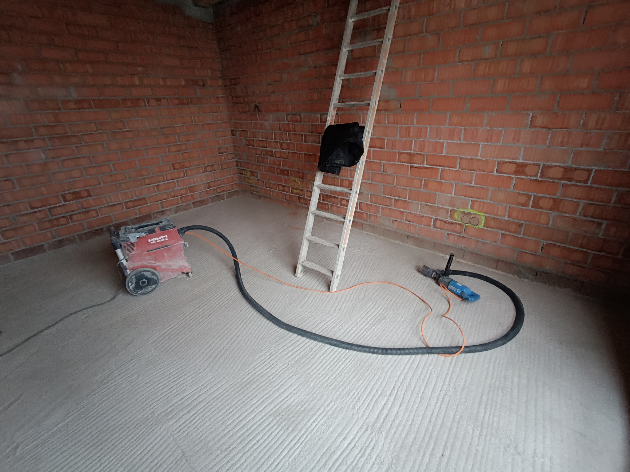 Wnętrze w trakcie prac: czerwona cegła, szara wylewka z bruzdami, drabina oparta o ścianę, odkurzacz przemysłowy Hilti i wiertarka z podłączonym odsysaniem pyłu.