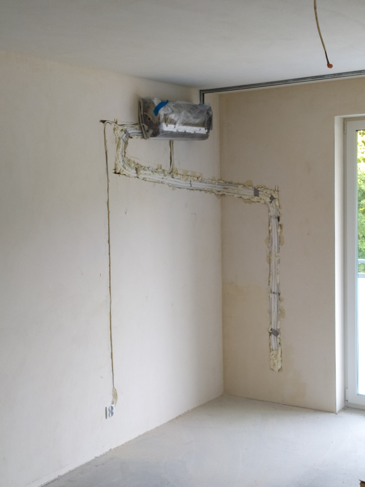 Białe kable elektryczne ułożone na ścianie w surowym wnętrzu, zabezpieczone pianką montażową, widoczne gniazdko. Prace instalacyjne w trakcie realizacji.