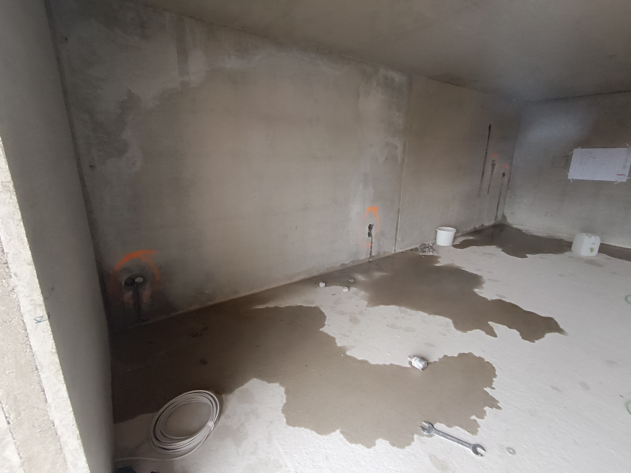 Surowy stan wnętrza z widocznymi instalacjami elektrycznymi oznaczonymi pomarańczowymi okręgami na ścianie, przewody i narzędzia na mokrej podłodze.