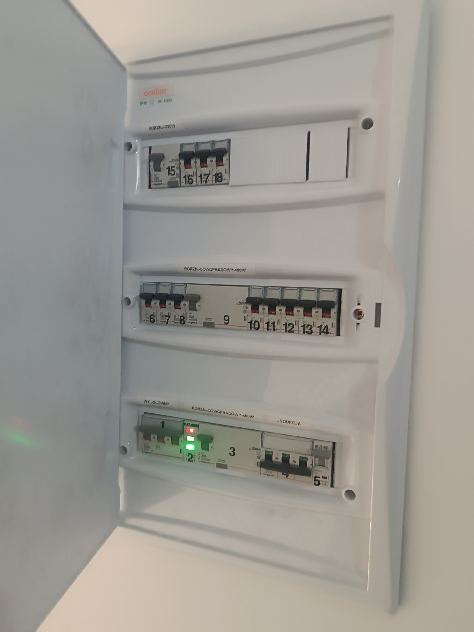 Otwarta, biała skrzynka rozdzielcza z bezpiecznikami. Widoczne oznaczenia i numery. Niektóre włączniki są w pozycji ON, inne OFF. Minimalistyczny design.