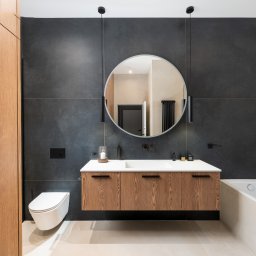 Ewa Stankiewicz EST Interior - Nowoczesna łazienka z drewnianą szafką pod umywalką, okrągłym lustrem i czarnymi płytkami. Minimalistyczny design z wiszącą toaletą i subtelnym oświetleniem.