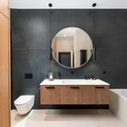 Nowoczesna łazienka z drewnianą szafką pod umywalką, okrągłym lustrem i czarnymi płytkami. Minimalistyczny design z wiszącą toaletą i subtelnym oświetleniem.