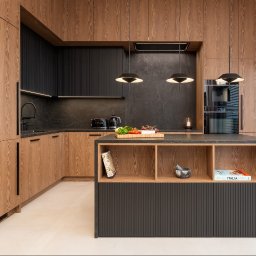 Ewa Stankiewicz EST Interior - Nowoczesna kuchnia z drewnianymi frontami i czarnym blatem. Wyspa kuchenna z otwartymi półkami. Minimalistyczne lampy wiszące. Duże okno z żaluzjami.