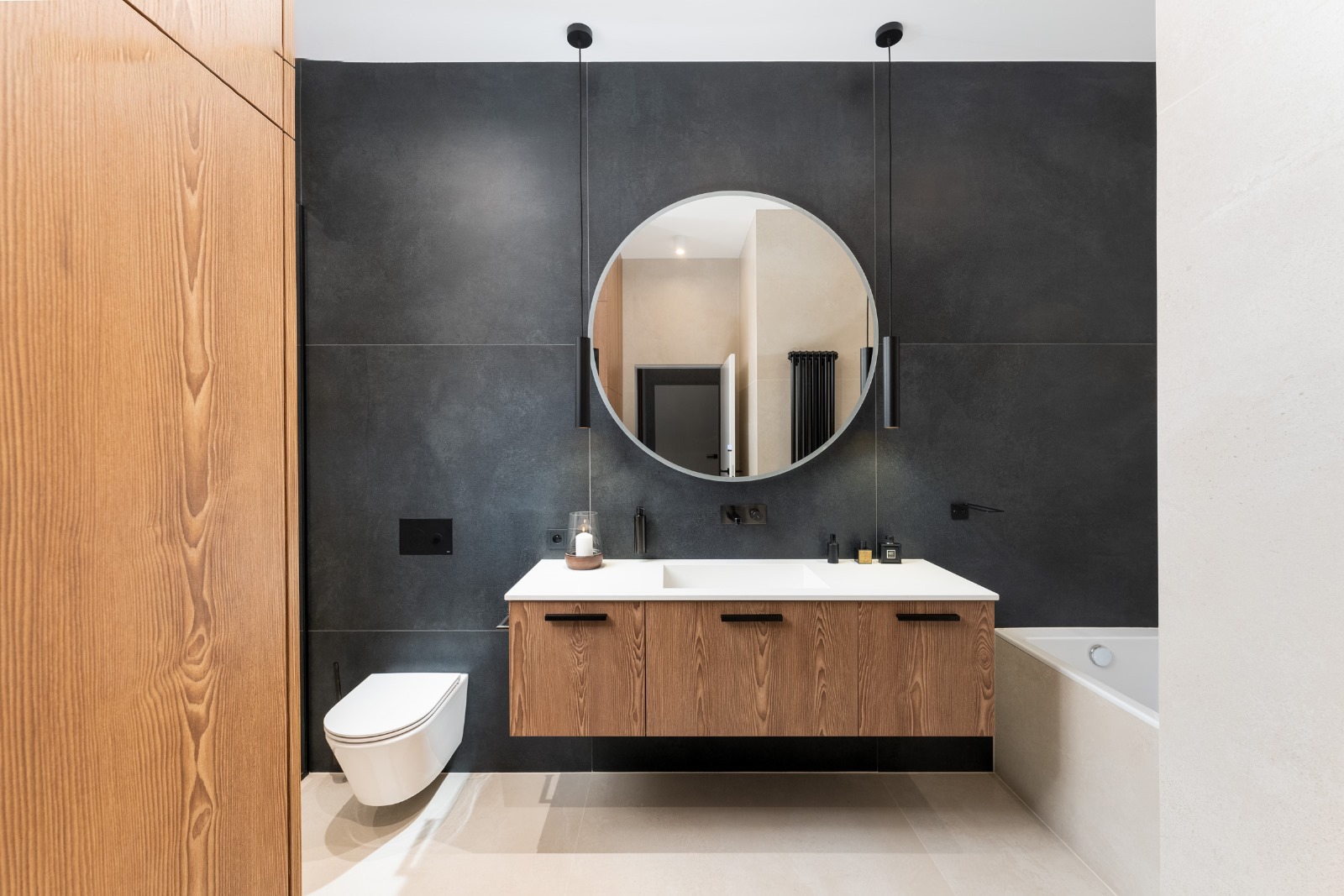 Nowoczesna łazienka z drewnianą szafką pod umywalką, okrągłym lustrem i czarnymi płytkami. Minimalistyczny design z wiszącą toaletą i subtelnym oświetleniem.