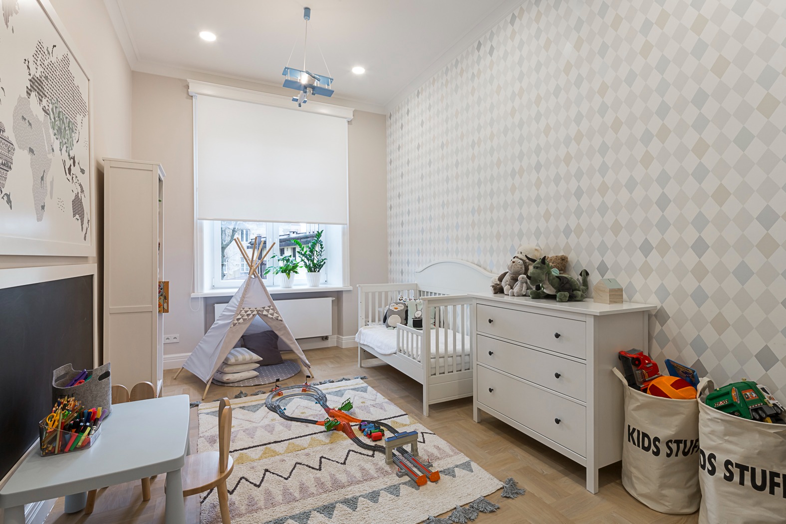 Jasny pokój dziecięcy z białymi meblami, tipi, łóżeczkiem i dywanem w geometryczne wzory. Dekoracje w postaci mapy świata i lampy w kształcie samolotu.