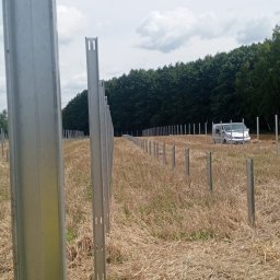 Helio Ground - Metalowe słupki ogrodzeniowe w rzędzie na polu, widoczny samochód dostawczy w tle. Etap montażu ogrodzenia panelowego na tle lasu.