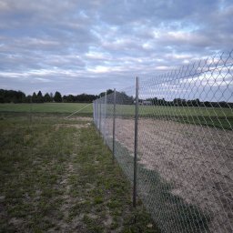 Helio Ground - Długi, metalowy płot z siatki na granicy działki, widok z perspektywy ziemi, pochmurne niebo w tle. Słupki ogrodzeniowe równomiernie rozstawione.