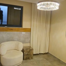 Invest-Bud - Beżowe wnętrze pokoju z okrągłym lustrem odbijającym fotel, obok okno z sylwetką osoby, kryształowy żyrandol i zasłony w ciepłej tonacji.