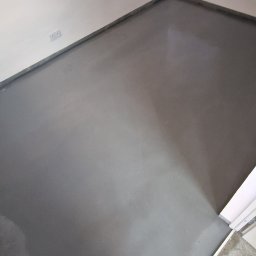 Invest-Bud - Świeżo wylana, samopoziomująca wylewka podłogowa w rogu pokoju, gotowa do dalszych prac wykończeniowych. Widoczny fragment progu i stopy osoby fotografującej.