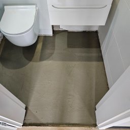 Invest-Bud - Wyrównana podłoga w łazience przed położeniem płytek. Widoczny fragment toalety i szafki pod umywalkę. Przygotowanie do wykończenia podłogi.