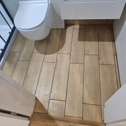 Invest-Bud - Wykończona łazienka z toaletą, szafką umywalkową i kabiną prysznicową. Na podłodze ułożone płytki imitujące drewno. Widok z góry, z otwartych drzwi.