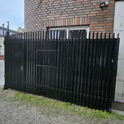 Czarny, metalowy płot z bramką na tle ceglanej ściany budynku z oknem. Widoczne kamienie i trawa u podstawy ogrodzenia.