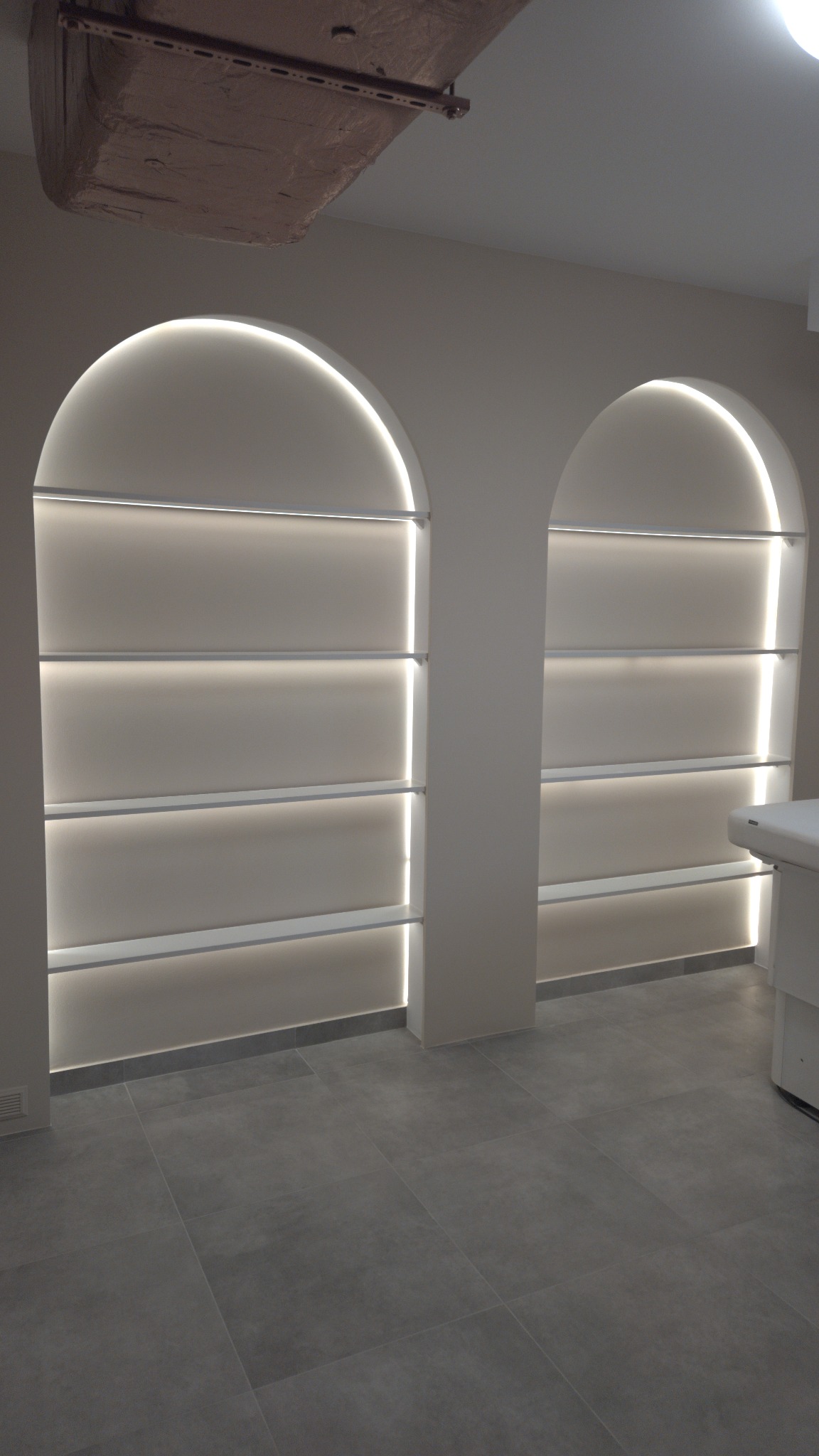 Dwie wnęki w kształcie łuków z podświetlanymi półkami w odcieniach beżu. Na suficie widoczny element dekoracyjny. Szare płytki na podłodze. Minimalistyczny design.