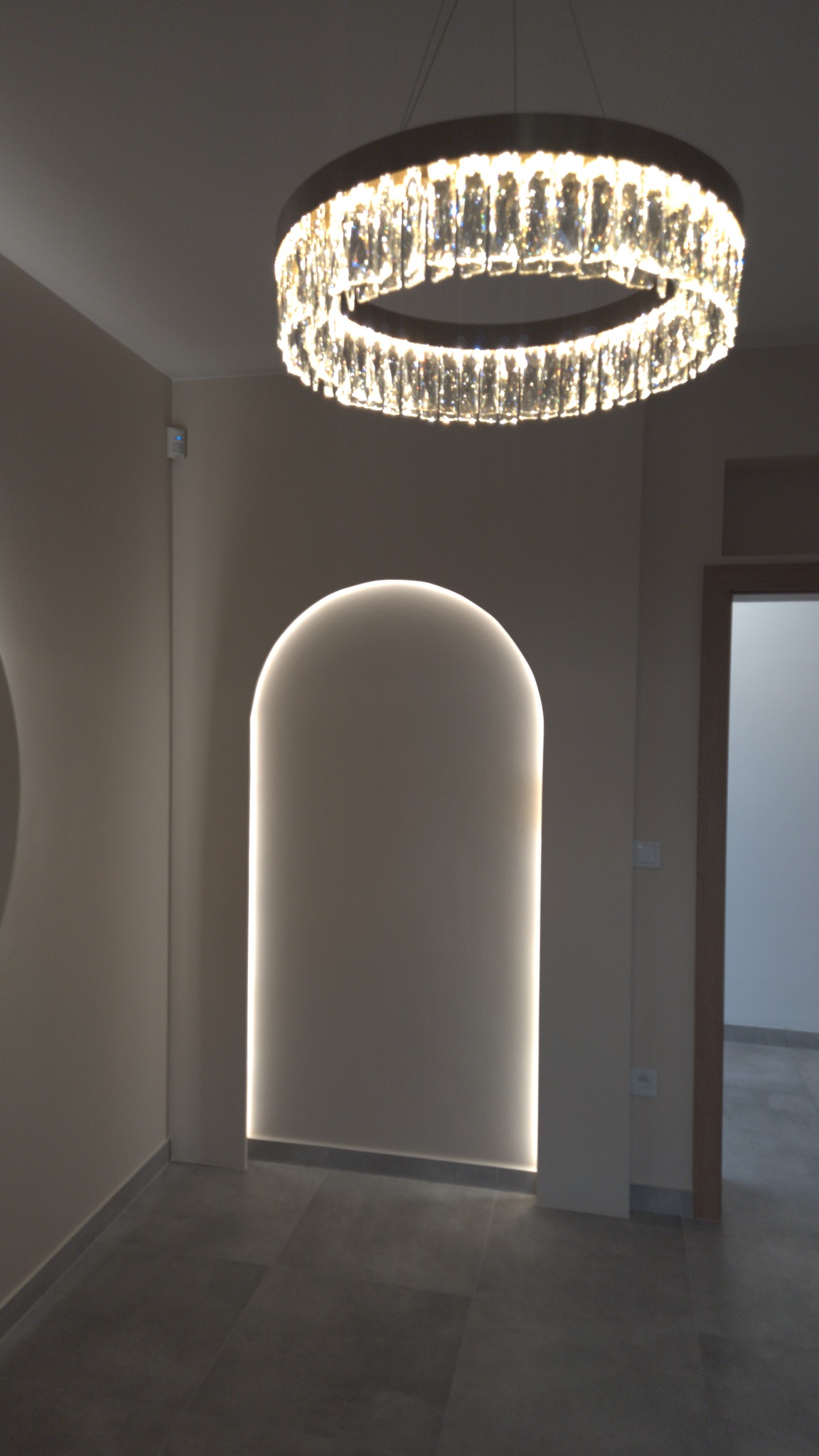 Nowoczesny hol z okrągłym żyrandolem kryształowym i wnęką z podświetleniem LED. Minimalistyczny design w neutralnej kolorystyce z szarymi płytkami podłogowymi.