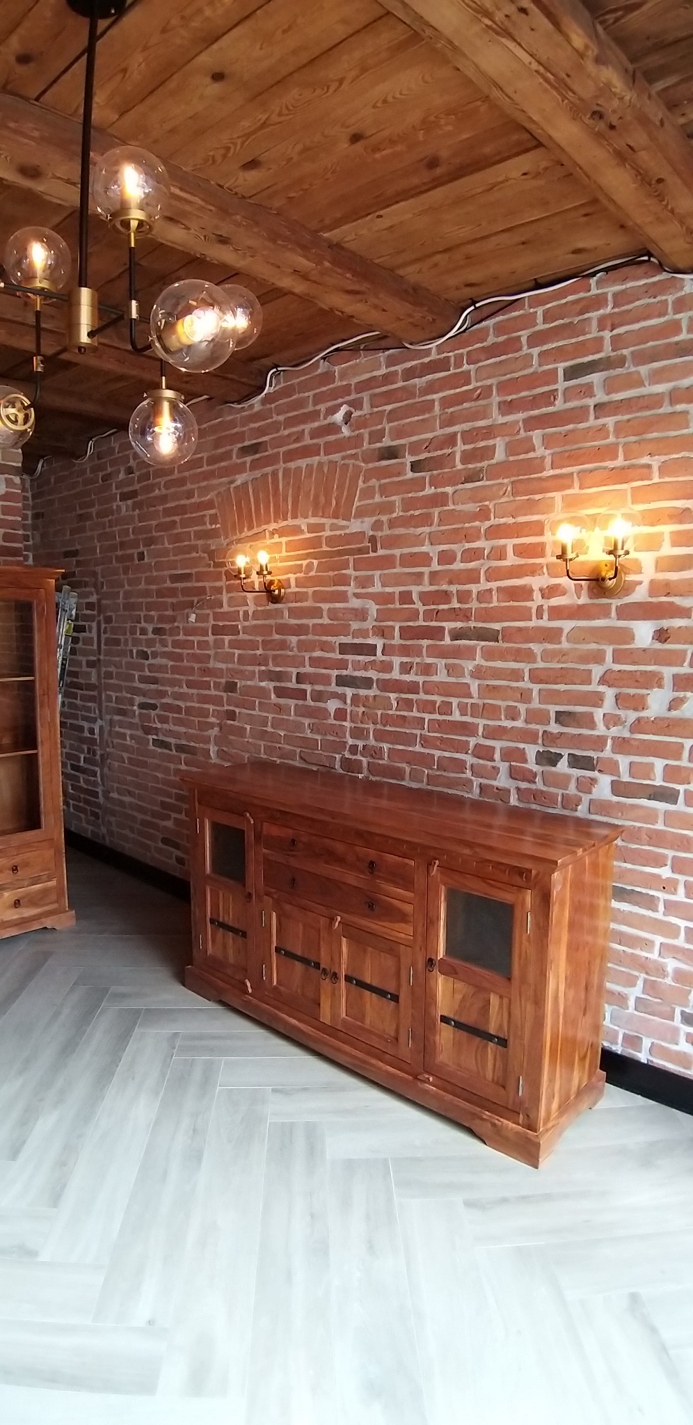 Rustykalne wnętrze: drewniany sufit, ceglana ściana z oświetleniem, komoda i witryna z litego drewna. Parkiet ułożony w jodełkę. Nowoczesna lampa sufitowa.