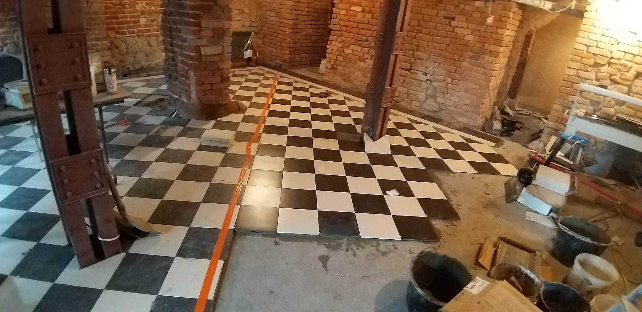 Układanie czarno-białej podłogi w szachownicę w pomieszczeniu z ceglanymi ścianami. Widoczne narzędzia i materiały budowlane. Prace wykończeniowe w trakcie.